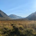 Breathtaking view of the Scottish Highlands with expansive fields and towering mountains under a clear blue sky.